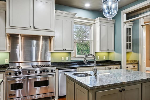 kitchen featuring a center island with sink, dark stone counters, range hood, appliances with stainless steel finishes, and a sink