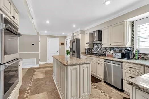 kitchen featuring light stone countertops, a kitchen island, stainless steel appliances, decorative backsplash, and wall chimney range hood