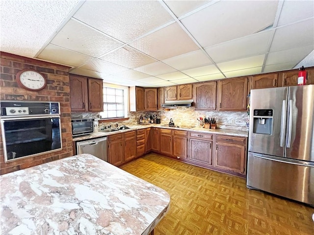 kitchen featuring a sink, decorative backsplash, light countertops, under cabinet range hood, and appliances with stainless steel finishes