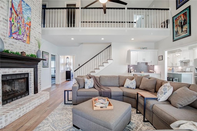 living area featuring a ceiling fan, a high ceiling, light wood-style floors, a brick fireplace, and stairs