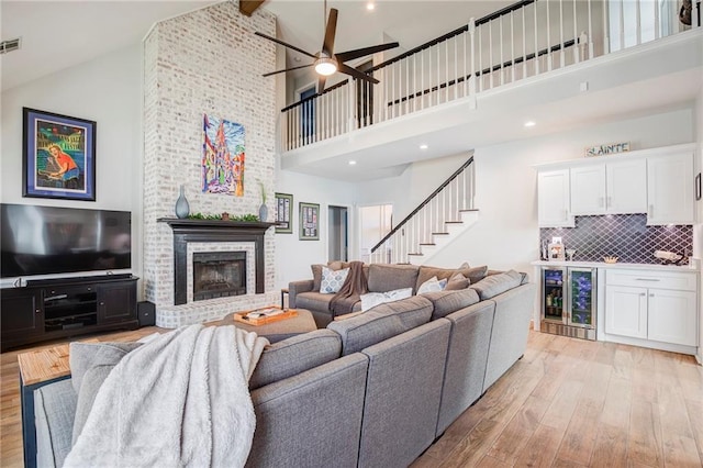 living area with high vaulted ceiling, light wood-style flooring, beverage cooler, a brick fireplace, and stairs