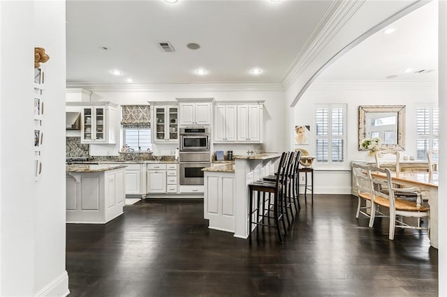 kitchen featuring glass insert cabinets, a breakfast bar area, arched walkways, white cabinetry, and wall chimney exhaust hood