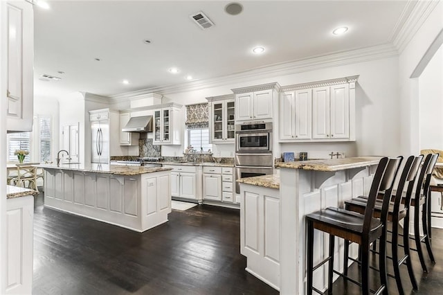 kitchen featuring visible vents, a breakfast bar, dark wood-type flooring, built in refrigerator, and wall chimney range hood