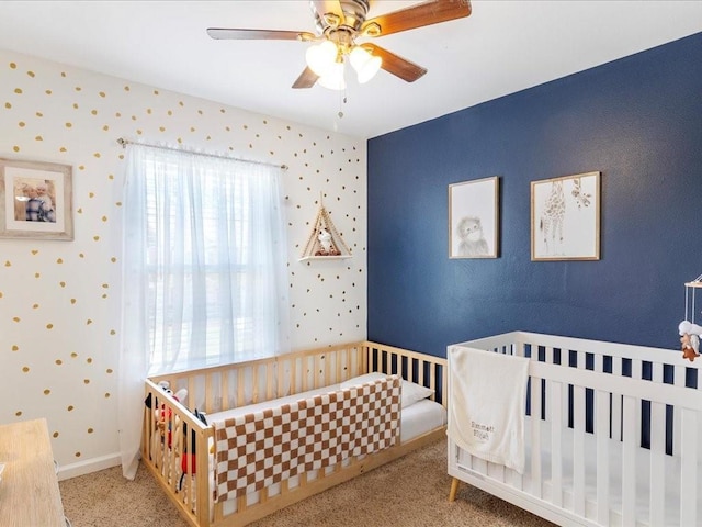 carpeted bedroom with a crib, wallpapered walls, baseboards, and a ceiling fan