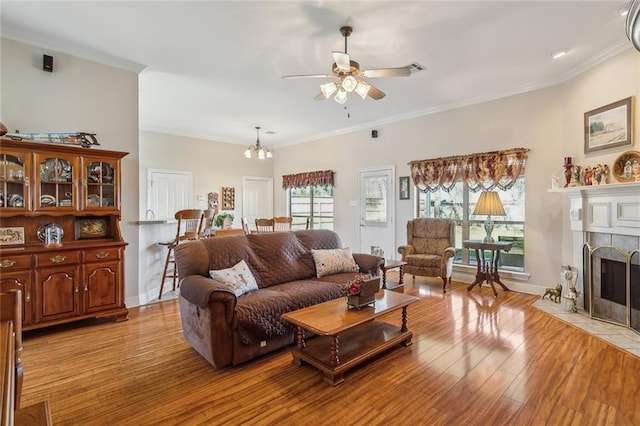 living area featuring ceiling fan with notable chandelier, light wood-style flooring, a tiled fireplace, and crown molding