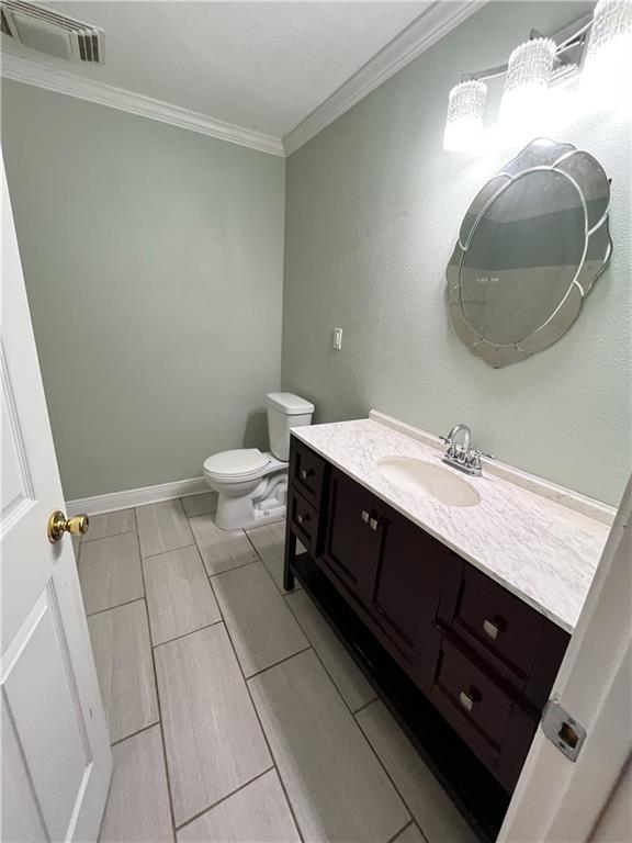 half bath with visible vents, toilet, ornamental molding, baseboards, and vanity