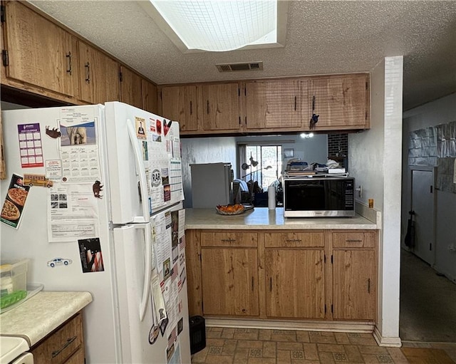 kitchen featuring a textured ceiling, freestanding refrigerator, brown cabinetry, light countertops, and brick floor