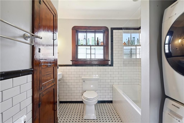 full bath featuring walk in shower, tile walls, stacked washer / dryer, toilet, and a bathtub