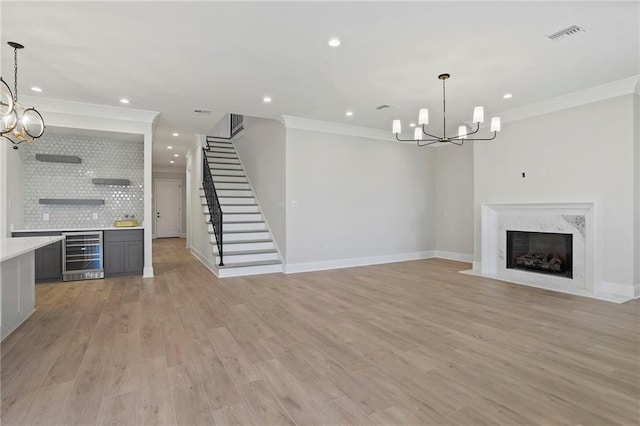 unfurnished living room with light wood finished floors, a premium fireplace, an inviting chandelier, wine cooler, and crown molding