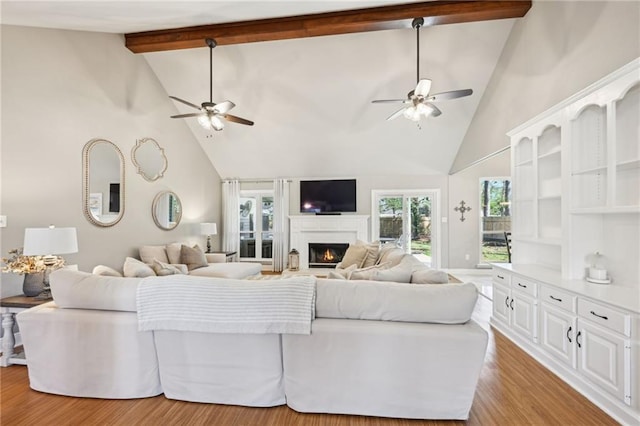 living room featuring a lit fireplace, beamed ceiling, light wood-style floors, and ceiling fan