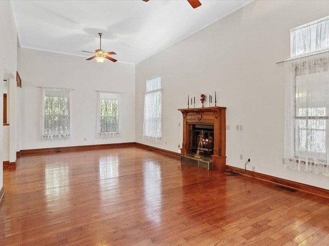 unfurnished living room featuring baseboards, hardwood / wood-style floors, a warm lit fireplace, high vaulted ceiling, and a ceiling fan