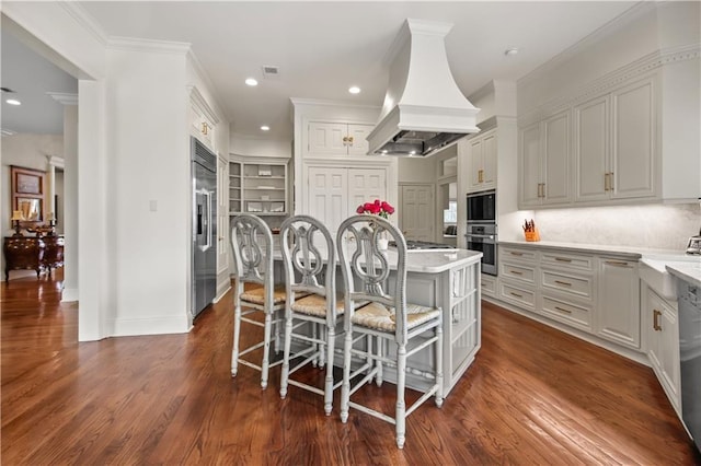 kitchen featuring premium range hood, a center island with sink, a breakfast bar, dark wood-style flooring, and built in appliances