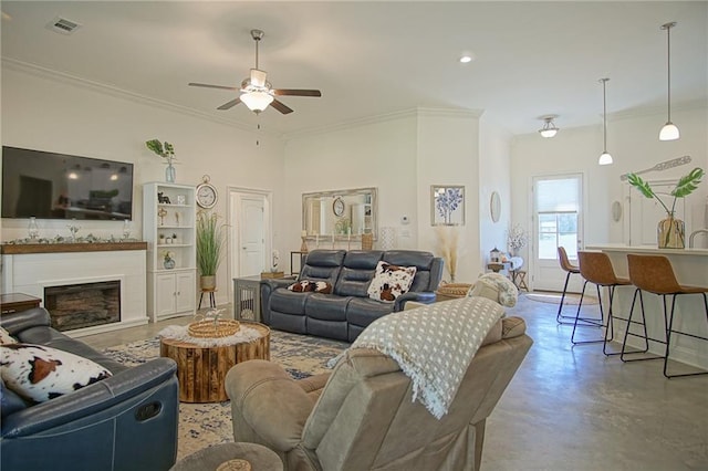 living area featuring finished concrete flooring, a ceiling fan, visible vents, ornamental molding, and a glass covered fireplace