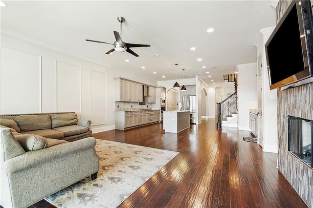 living room featuring stairway, a fireplace, arched walkways, ornamental molding, and ceiling fan