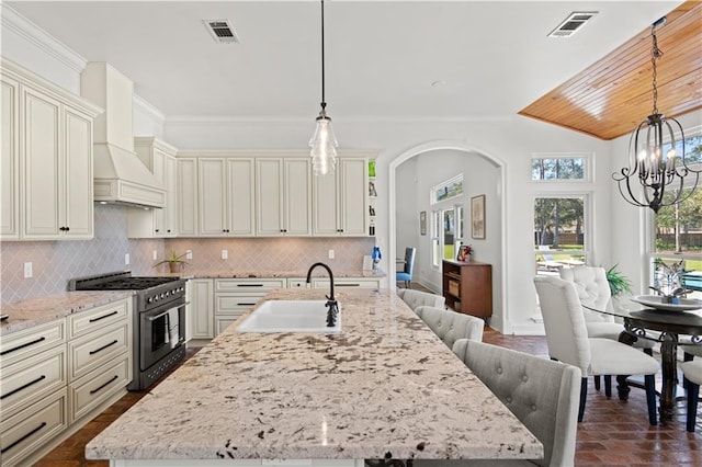 kitchen with a notable chandelier, high end stove, a sink, backsplash, and custom exhaust hood