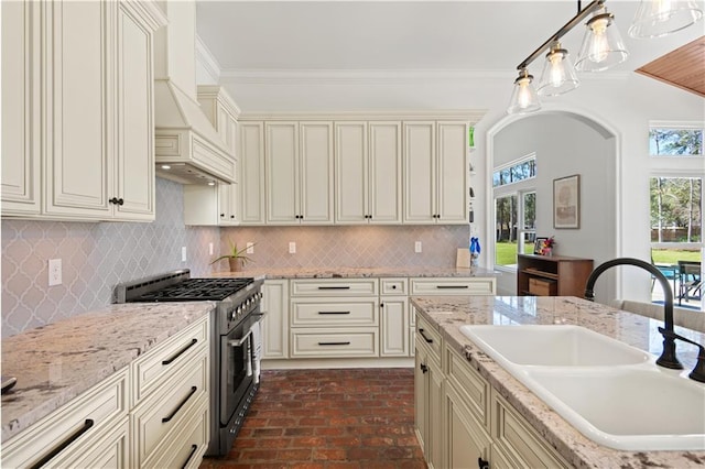 kitchen with custom exhaust hood, brick floor, a sink, stainless steel range, and cream cabinetry