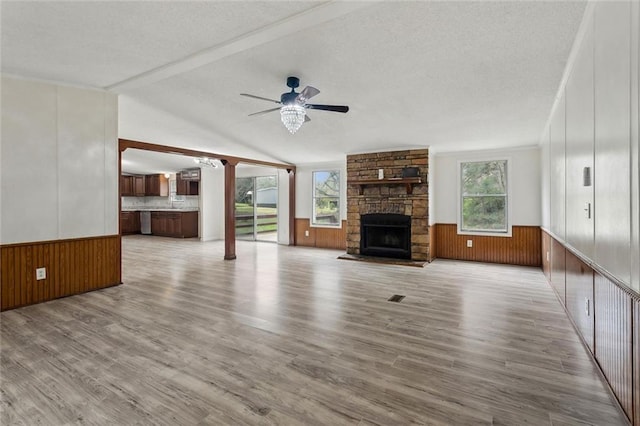 unfurnished living room with vaulted ceiling with beams, ceiling fan, a wainscoted wall, a stone fireplace, and a textured ceiling