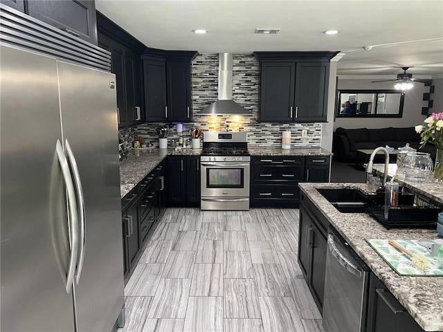 kitchen featuring a sink, backsplash, appliances with stainless steel finishes, wall chimney exhaust hood, and dark cabinets