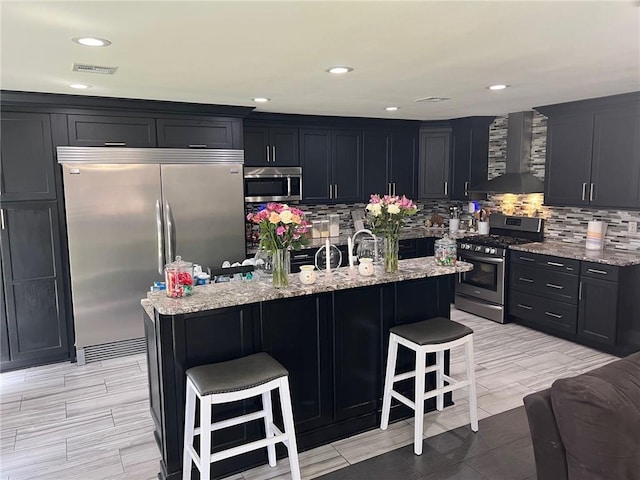 kitchen featuring visible vents, wall chimney range hood, a breakfast bar area, light stone counters, and appliances with stainless steel finishes