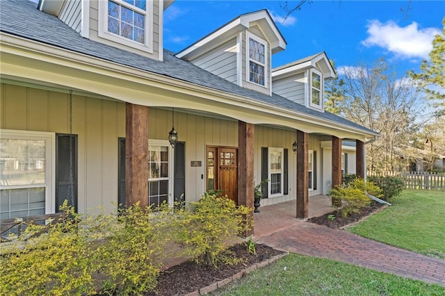 view of exterior entry with a porch, roof with shingles, a yard, and fence