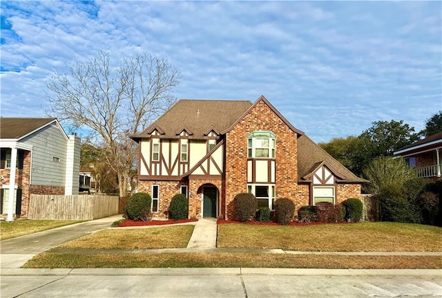 tudor house with brick siding, a front yard, roof with shingles, and fence