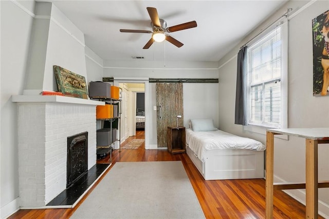 bedroom featuring ceiling fan, baseboards, a barn door, a fireplace, and wood finished floors