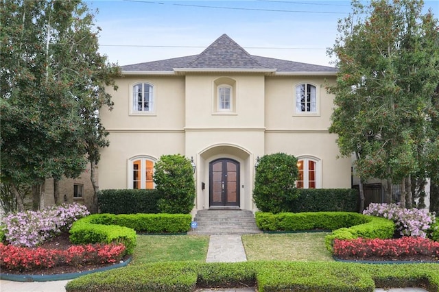 french country inspired facade featuring stucco siding, a shingled roof, a front yard, and french doors