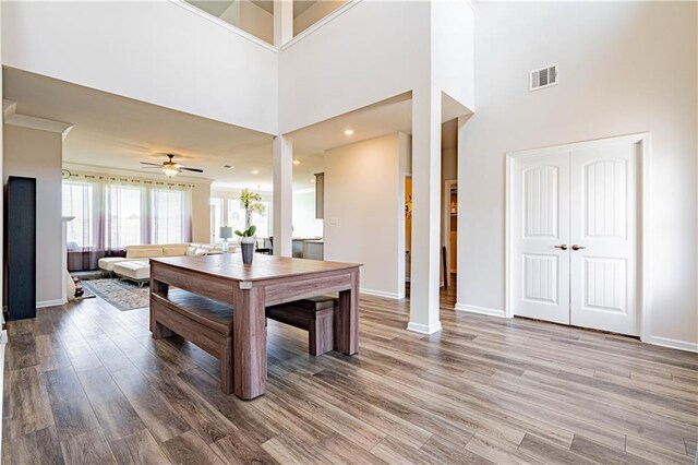 dining space featuring wood finished floors, baseboards, visible vents, ceiling fan, and a towering ceiling