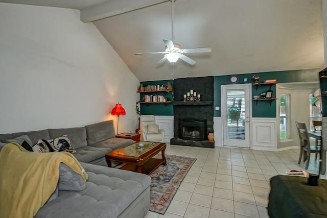 living room with a fireplace, vaulted ceiling with beams, a wainscoted wall, a ceiling fan, and light tile patterned floors