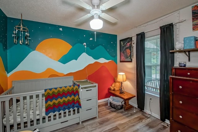 bedroom featuring a nursery area, a textured ceiling, a ceiling fan, baseboards, and wood finished floors
