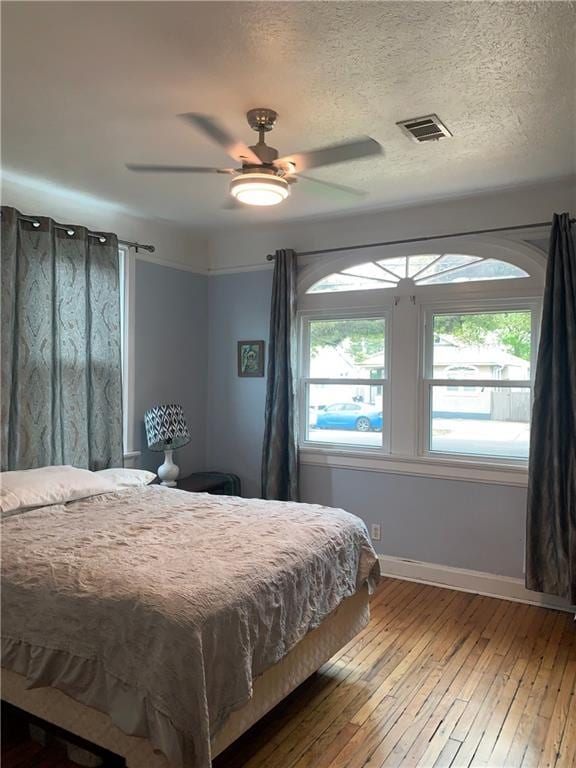 bedroom with baseboards, a textured ceiling, hardwood / wood-style flooring, visible vents, and a ceiling fan