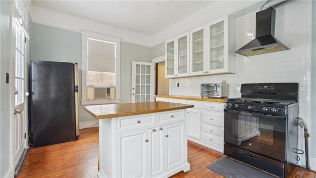 kitchen with black gas stove, wood finished floors, white cabinetry, wall chimney range hood, and freestanding refrigerator