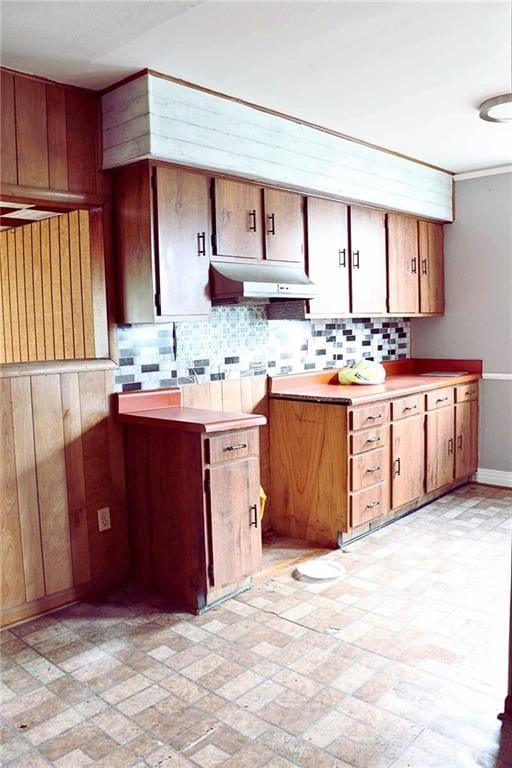 kitchen with light countertops, baseboards, under cabinet range hood, backsplash, and brown cabinets