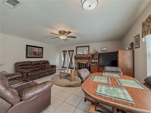 living room featuring a textured ceiling, a ceiling fan, a glass covered fireplace, visible vents, and light tile patterned flooring