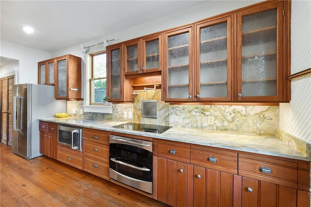 kitchen with light wood-style floors, brown cabinetry, light stone countertops, backsplash, and stainless steel appliances