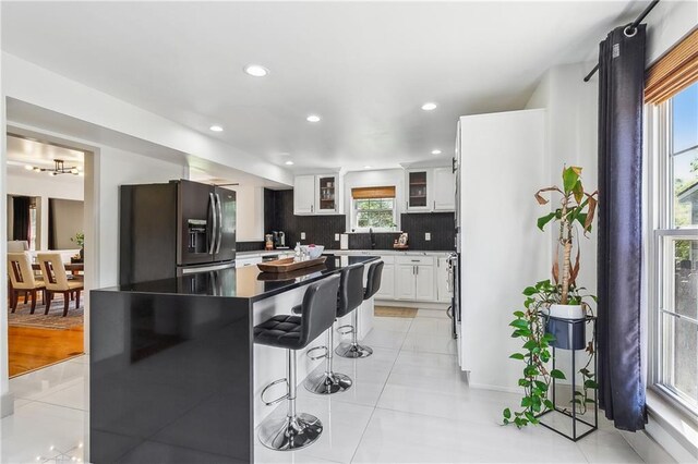 kitchen with tasteful backsplash, a breakfast bar, light tile patterned floors, white cabinets, and black refrigerator with ice dispenser