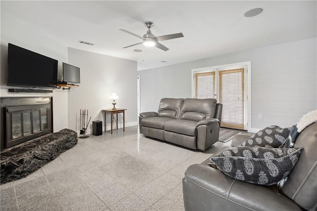 living room featuring a ceiling fan, visible vents, granite finish floor, baseboards, and a glass covered fireplace