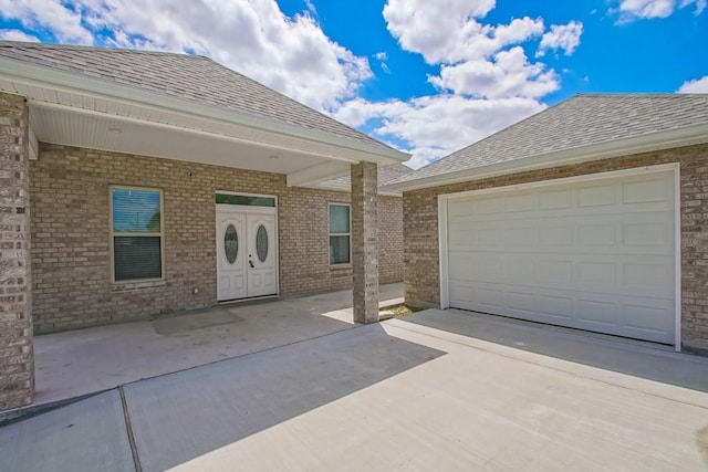 view of exterior entry featuring roof with shingles, a garage, concrete driveway, and brick siding