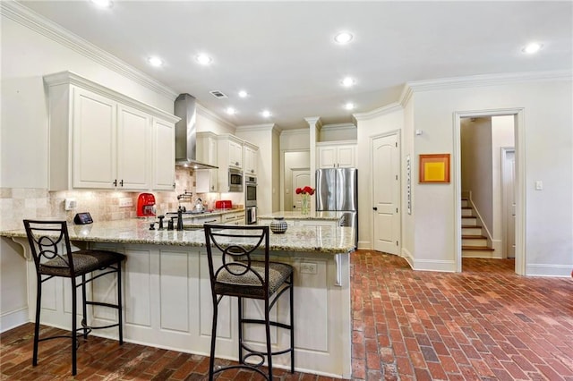 kitchen with wall chimney range hood, tasteful backsplash, brick floor, a peninsula, and visible vents