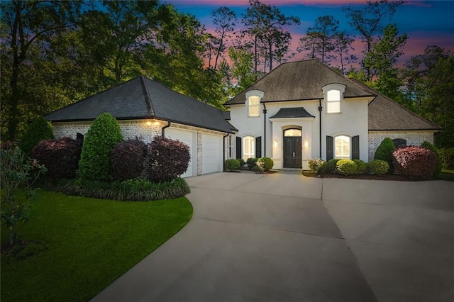 french provincial home featuring concrete driveway, a garage, brick siding, a lawn, and stucco siding