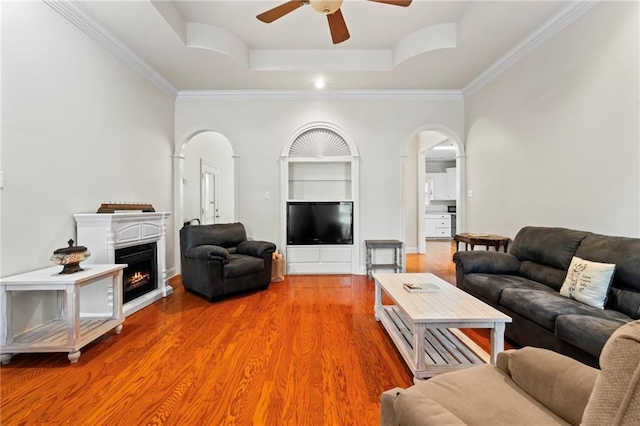 living room with a ceiling fan, arched walkways, a raised ceiling, light wood-style floors, and ornamental molding