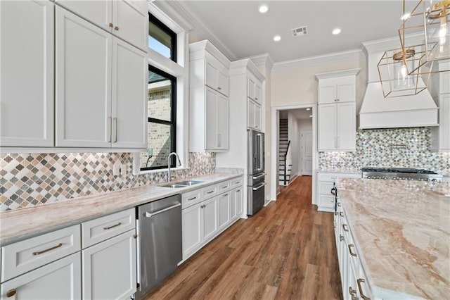 kitchen with a sink, stainless steel appliances, custom range hood, white cabinetry, and ornamental molding