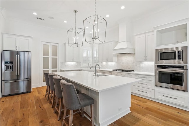 kitchen featuring a sink, appliances with stainless steel finishes, light countertops, premium range hood, and a center island with sink