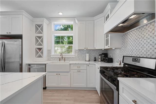 kitchen with custom exhaust hood, appliances with stainless steel finishes, white cabinets, a sink, and light wood-type flooring