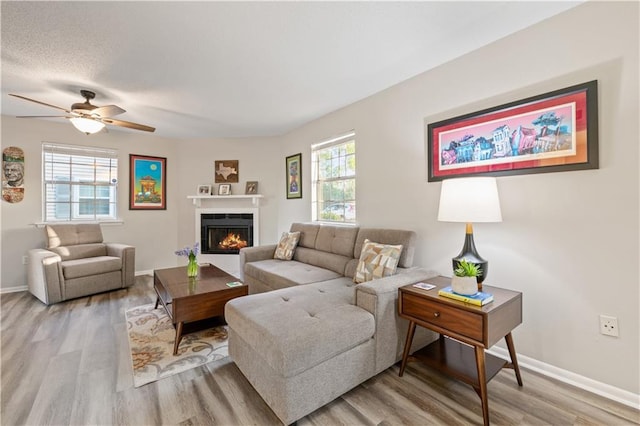 living area featuring baseboards, light wood-type flooring, ceiling fan, and a lit fireplace