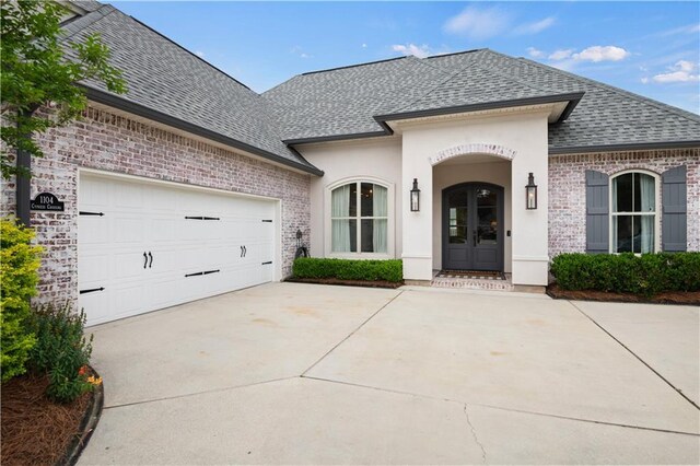 french country inspired facade with concrete driveway, a shingled roof, french doors, brick siding, and a garage