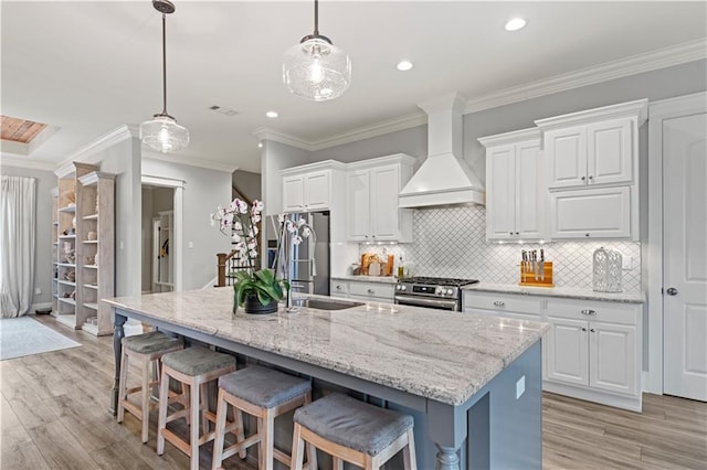 kitchen with a breakfast bar area, appliances with stainless steel finishes, light wood-style floors, custom exhaust hood, and an island with sink