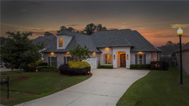french country style house with concrete driveway, roof with shingles, a lawn, brick siding, and a garage