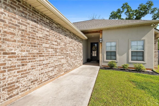 view of exterior entry with brick siding, a shingled roof, a lawn, and stucco siding