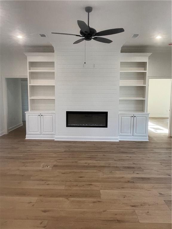 unfurnished living room with a large fireplace, ceiling fan, built in features, and wood finished floors